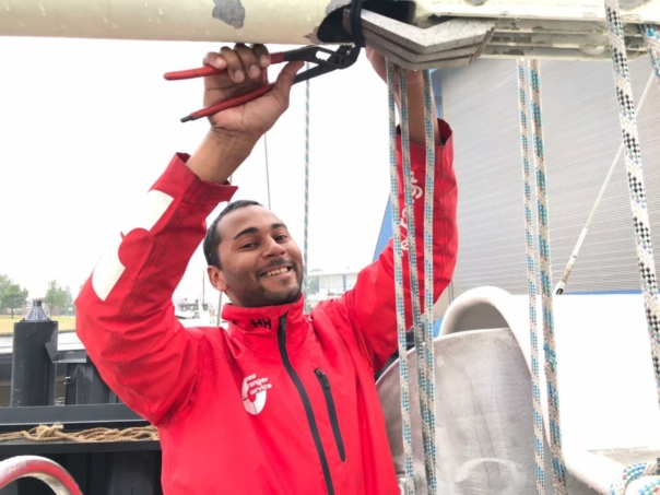 A male Sea Ranger in a red Sea Ranger jacket poses for the camera. The man has a pair of pliers in his hand and is fixing the rigging on the boat.