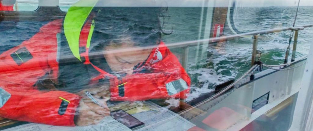 A Sea Ranger in uniform, sat at a desk on board a ship. The Sea Ranger is looking at their phone and making notes using pen and paper.