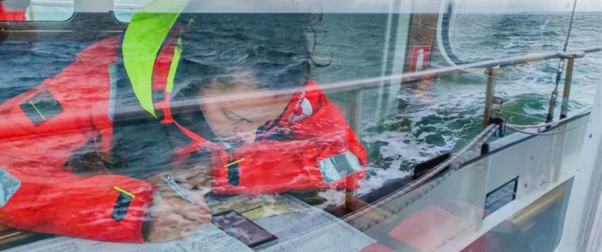 A Sea Ranger in uniform, sat at a desk on board a ship. The Sea Ranger is looking at their phone and making notes using pen and paper.