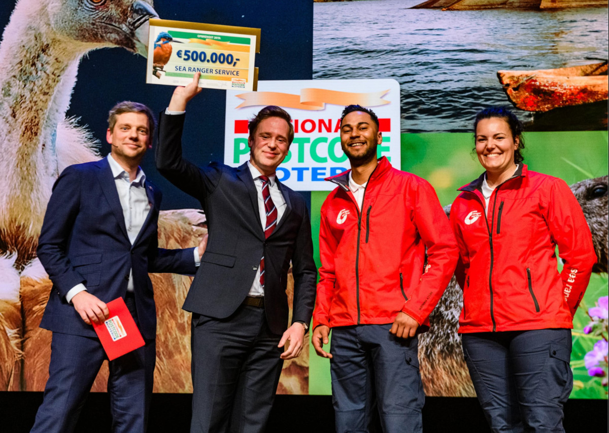 Sea Ranger Service founder Wietse van der Werf, two Sea Rangers in uniform, and Margriet Schreuders, Head of Charities at the Postcode Lottery are photographed on a stage. Wietse van der Werf holds a cheque for 500,000 euros above his head.