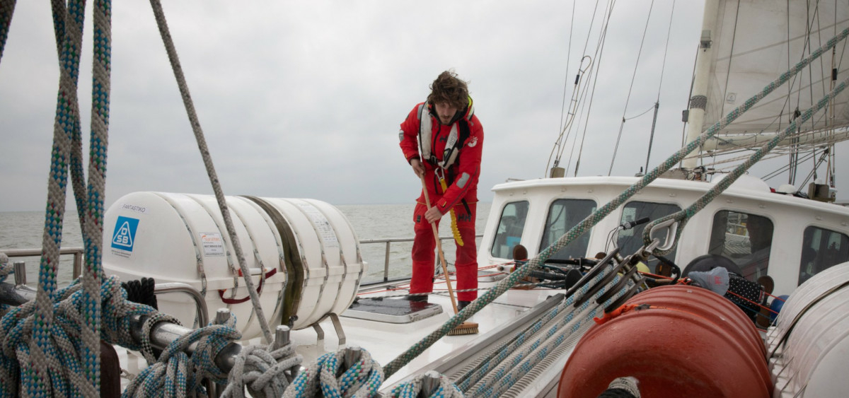 A lone Sea Ranger in uniform is on the deck of a ship, cleaning it with a long-handled brush. The sky and sea in the background are gray.