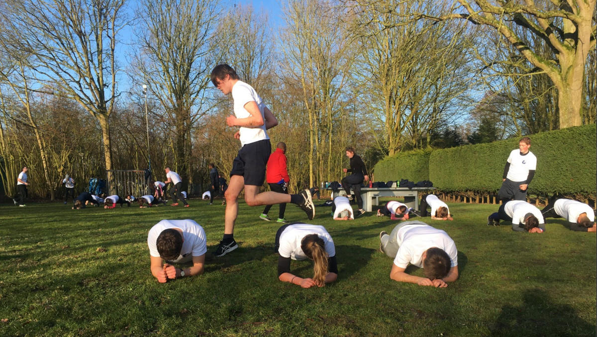 Bootcamp recruits compete in shuttle runs, hurdling over other recruits who are planking. The recruits are dressed in white t-shirts and dark shorts. It is a sunny spring day. The sky is clear, and the trees in the background have no leaves.