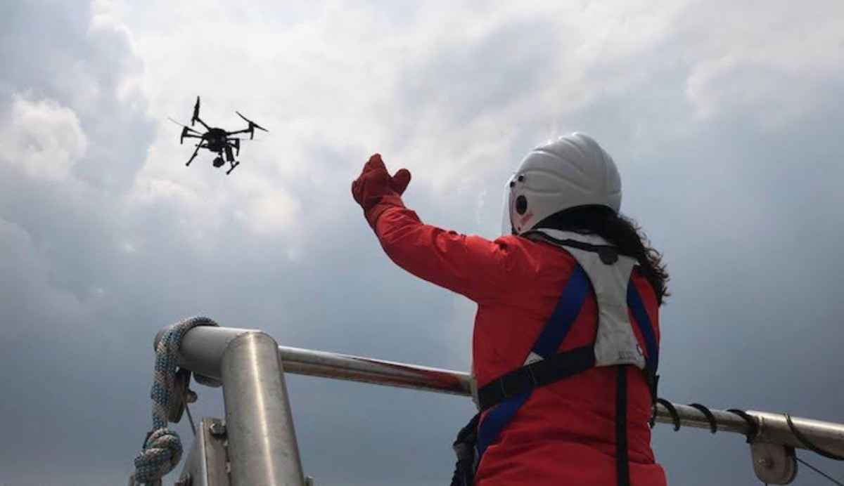 A Sea Ranger launches a drone into the sky, from a ship at sea, to carry out ocean conservation work. The Sea Ranger is in a red uniform, and the sky is bright.
