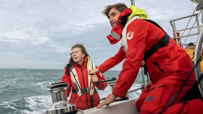 A male and female Sea Ranger, dressed in red Sea Ranger uniform, are sailing a ship. The ocean can be seen in the background.