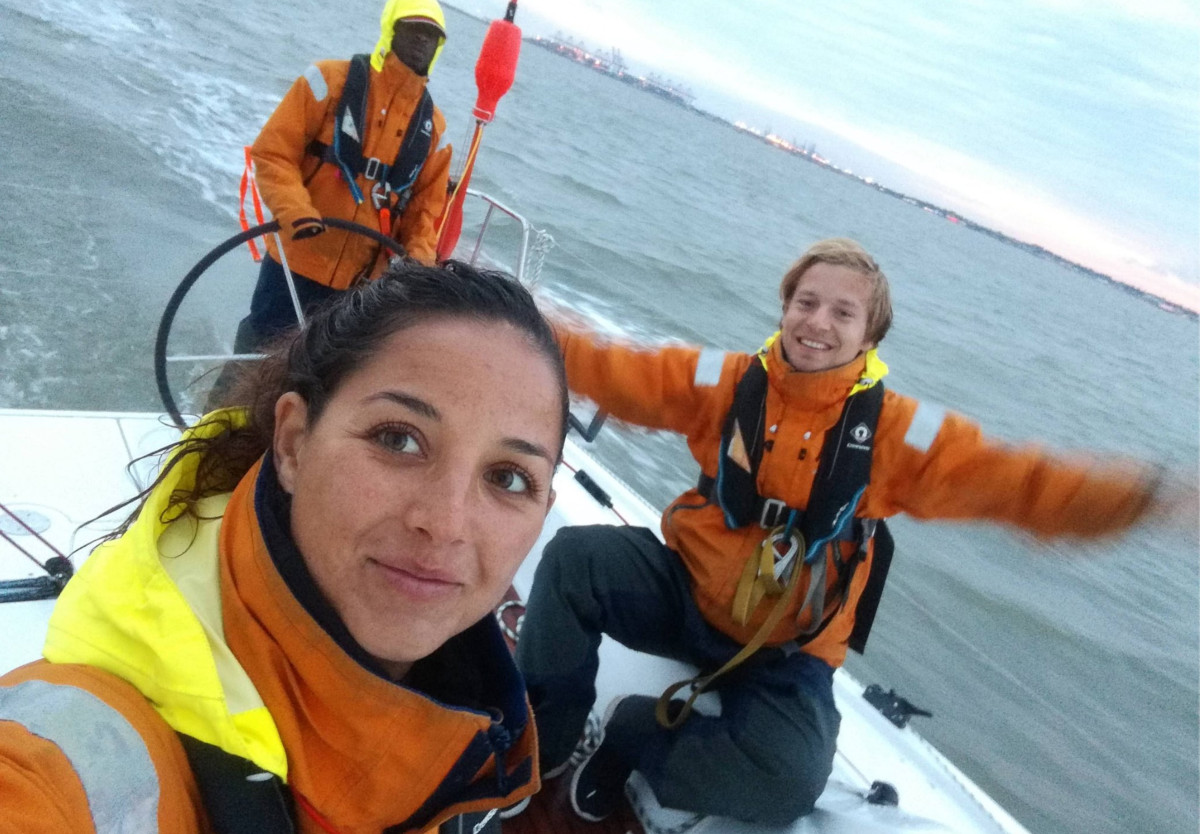 Three Sea Rangers at sea, in orange jackets, pose for a selfie. In the foreground, a woman is smiling. In the background, one of the male Sea Rangers has his arms spread wide and is smiling, while the other is steering the boat.