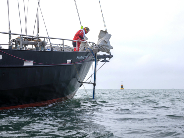A Sea Ranger dressed in a red uniform is looking over the side of a navy blue ship at the ocean, undertaking hydrographic surveying.